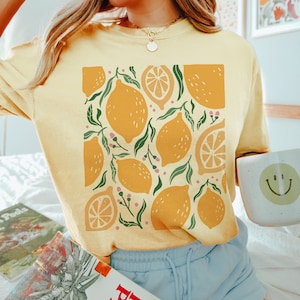 Puede incluir: Una camiseta amarilla con un estampado gr&aacute;fico de limones y hojas. Los limones son de varios tonos de amarillo y naranja. Las hojas son verdes. La camiseta la lleva puesta una persona con el pelo largo y rubio.