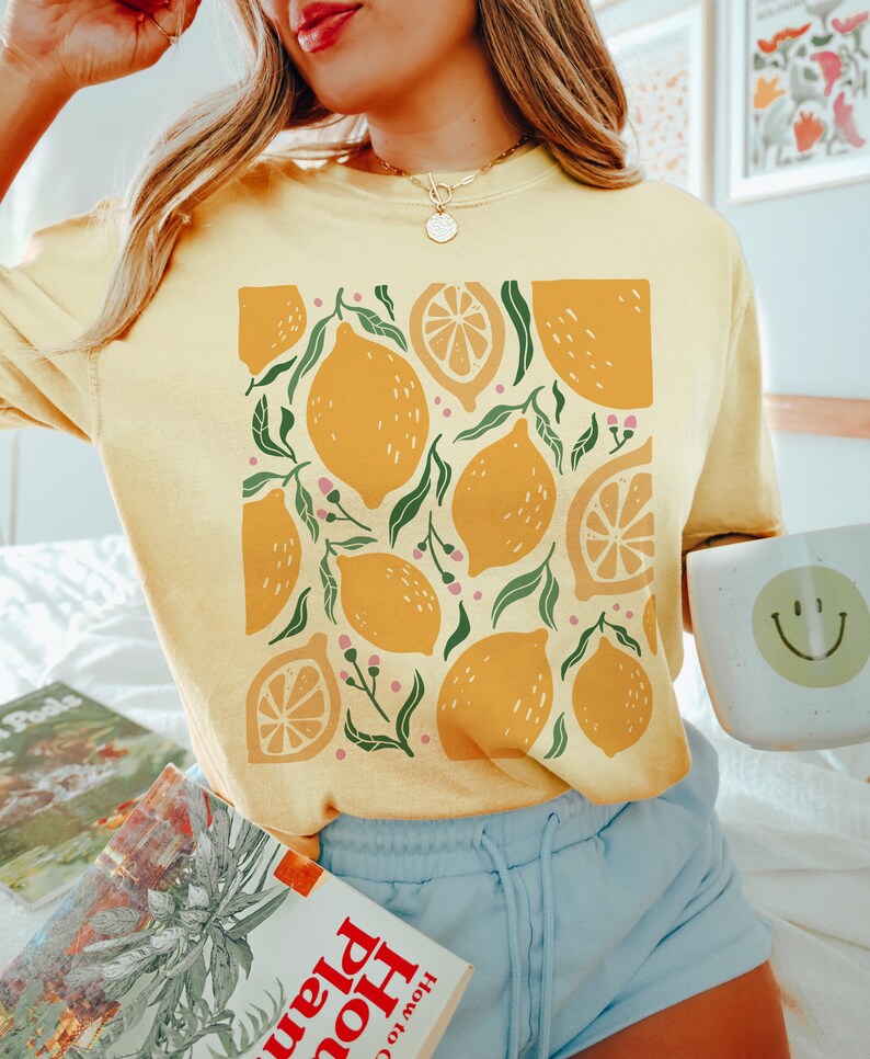 Puede incluir: Una camiseta amarilla con un estampado gr&aacute;fico de limones y hojas. Los limones son de varios tonos de amarillo y naranja. Las hojas son verdes. La camiseta la lleva puesta una persona con el pelo largo y rubio.