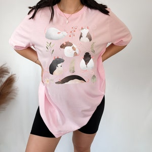 May include: A pink t-shirt with a graphic print of several rats in different poses. The rats are illustrated in a cartoon style with soft colors and are surrounded by leaves and flowers.