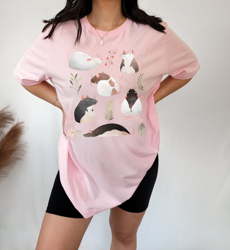 May include: A pink t-shirt with a graphic print of several rats in different poses. The rats are illustrated in a cartoon style with soft colors and are surrounded by leaves and flowers.