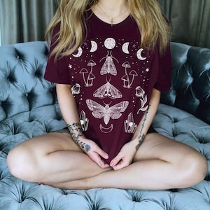 Puede incluir: Una camiseta color burdeos con un dise&ntilde;o gr&aacute;fico blanco que presenta polillas, setas y lunas crecientes. El dise&ntilde;o est&aacute; centrado en la camiseta e incluye el texto "the moon whispers to my soul".