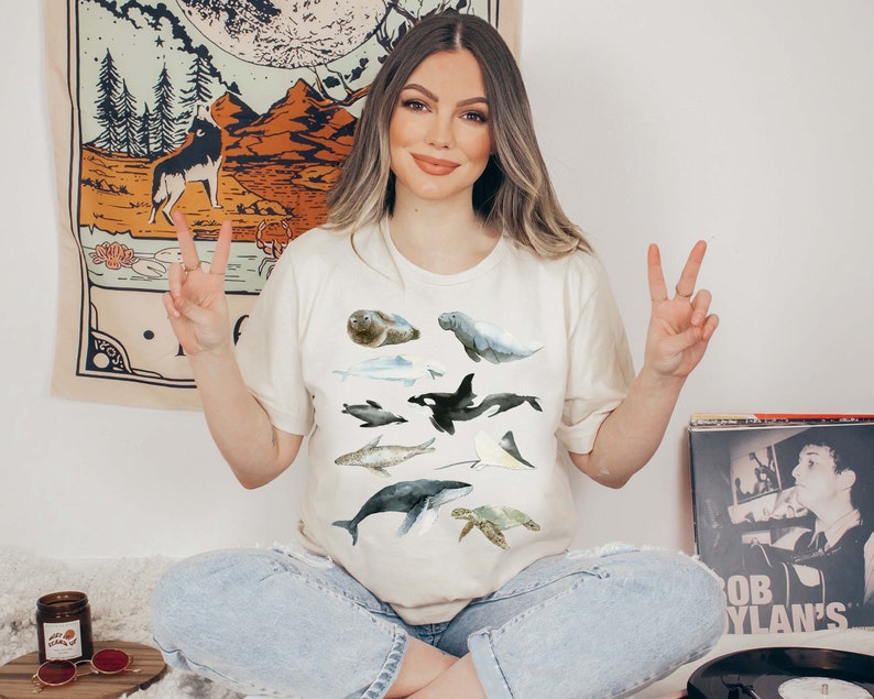 K&ouml;nnte beinhalten: Eine Frau tr&auml;gt ein cremefarbenes T-Shirt mit einem Aquarell-Design aus verschiedenen Meerestieren, darunter ein Wal, eine Meeresschildkr&ouml;te, ein Seekuh und eine Robbe. Die Frau sitzt mit gekreuzten Beinen und macht mit beiden H&auml;nden das Peace-Zeichen.