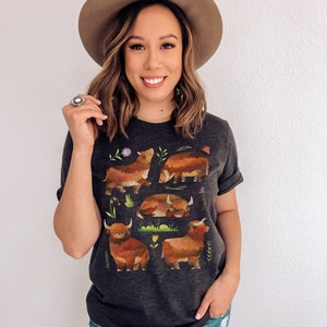 Puede incluir: Una camiseta gris oscuro con un estampado gr&aacute;fico de cinco vacas Highland marrones y naranjas con hierba verde y flores. Las vacas est&aacute;n en diferentes poses, incluyendo de pie, acostadas y mirando hacia un lado.