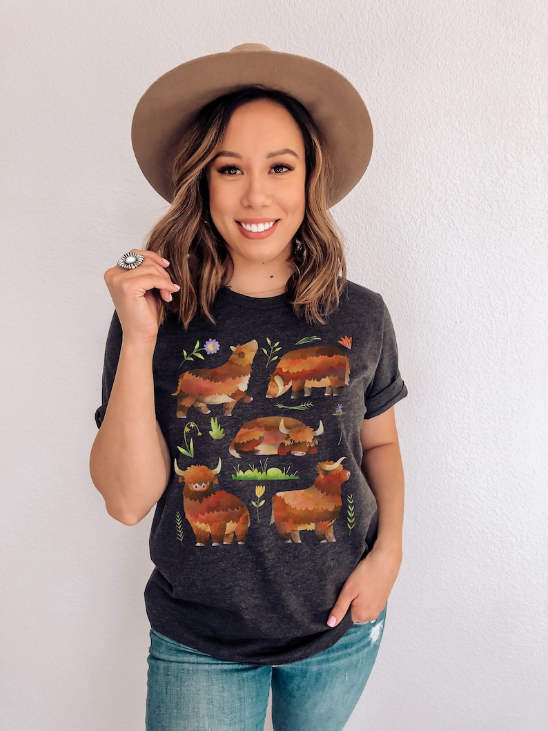 Puede incluir: Una camiseta gris oscuro con un estampado gr&aacute;fico de cinco vacas Highland marrones y naranjas con hierba verde y flores. Las vacas est&aacute;n en diferentes poses, incluyendo de pie, acostadas y mirando hacia un lado.