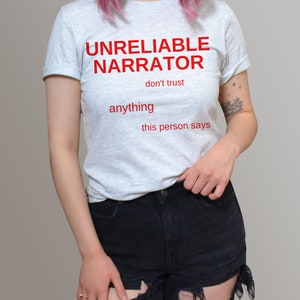 Puede incluir: Una camiseta gris claro con el texto "UNRELIABLE NARRATOR don't trust anything this person says" impreso en rojo.