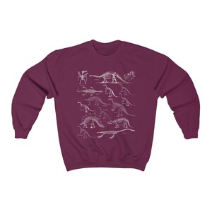 May include: A maroon sweatshirt with a white print of dinosaur skeletons. The print features a variety of dinosaurs, including a stegosaurus, a triceratops, and a brachiosaurus.