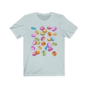 May include: A light blue t-shirt with a pattern of colorful macarons. The macarons are scattered randomly across the shirt.
