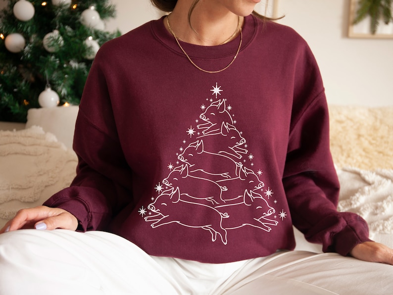 May include: A maroon sweatshirt featuring a white outline of a Christmas tree made of pigs. The pigs are stacked on top of each other in a pyramid shape.