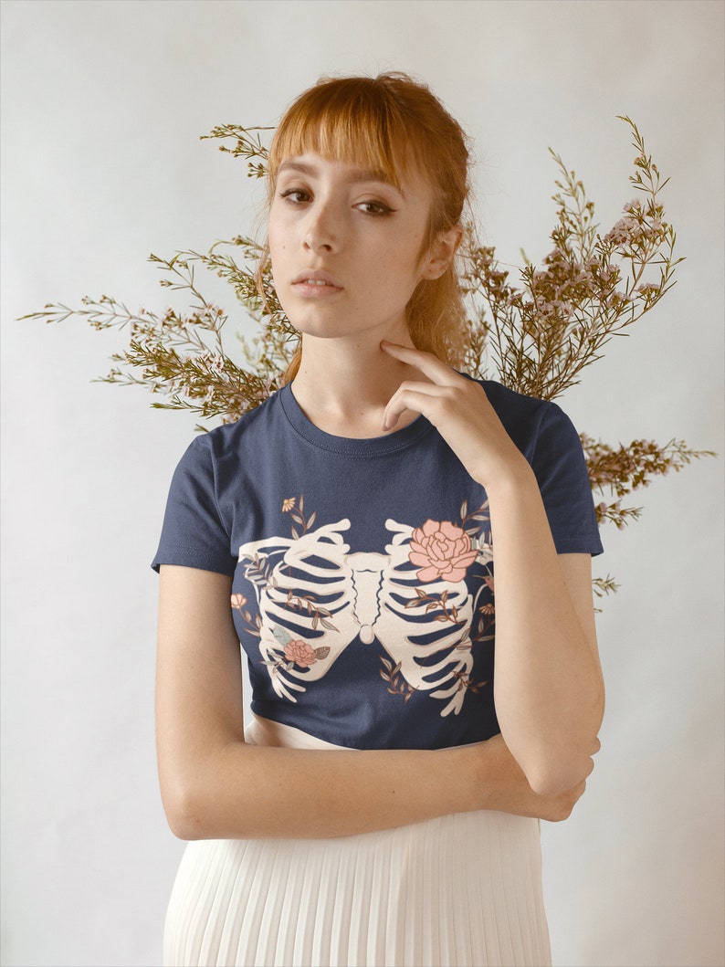 May include: A navy blue t-shirt with a white ribcage design and floral accents. The ribcage is surrounded by pink and white flowers and leaves.