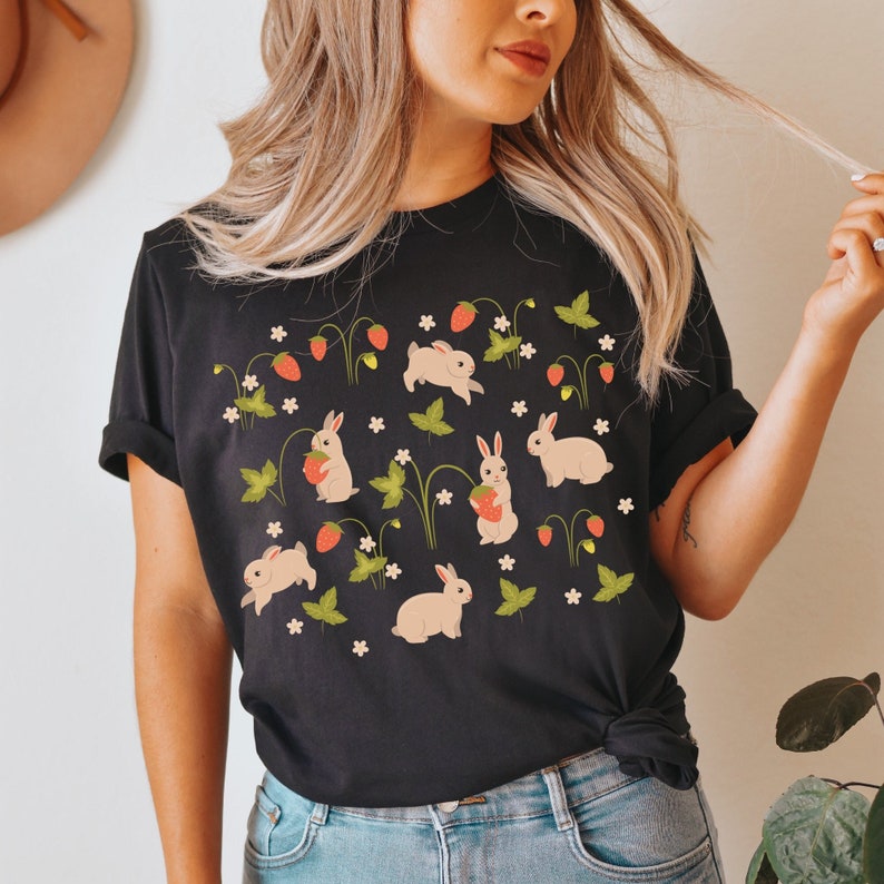 Op de afbeelding: Zwart T-shirt met een witte konijnen- en aardbeienprint. De print toont meerdere konijnen die aardbeien vasthouden en omringd zijn door groene bladeren en witte bloemen.