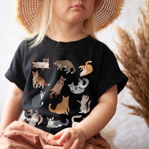 May include: A child wearing a black t-shirt with a print of various cats in different poses. The shirt has a crew neck and short sleeves. The child is wearing a straw hat and is sitting down.