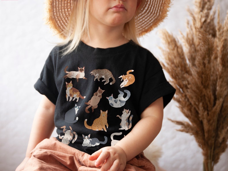 May include: A child wearing a black t-shirt with a print of various cats in different poses. The shirt has a crew neck and short sleeves. The child is wearing a straw hat and is sitting down.