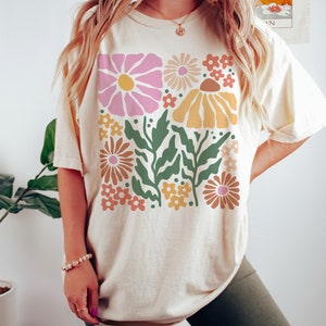 Puede incluir: Una camiseta de color crema con un estampado floral retro en tonos rosa, amarillo, naranja y verde. El estampado presenta flores y hojas estilizadas.