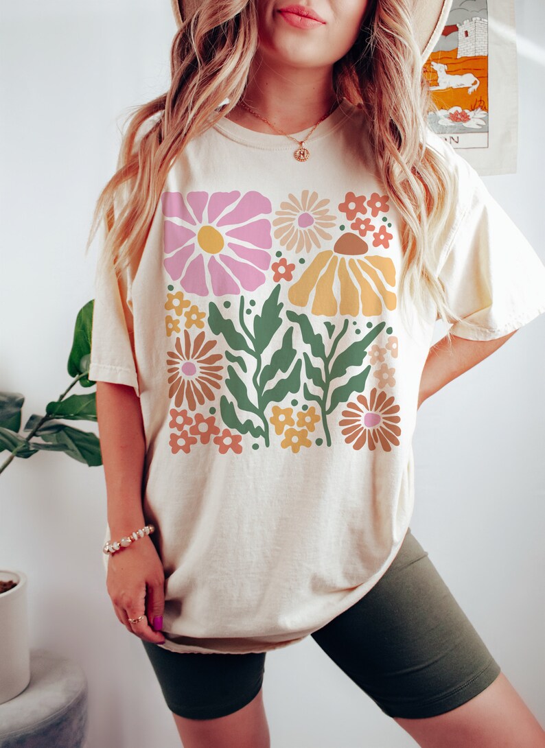Puede incluir: Una camiseta de color crema con un estampado floral retro en tonos rosa, amarillo, naranja y verde. El estampado presenta flores y hojas estilizadas.