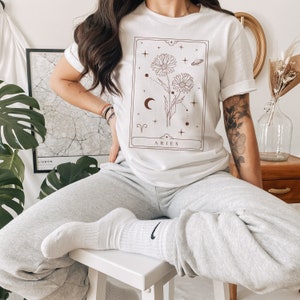 May include: White t-shirt with a brown graphic of a flower and constellation with the text "Aries".