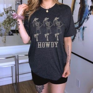 K&ouml;nnte beinhalten: Ein dunkelgraues T-Shirt mit einem Aufdruck von drei Skeletten, die Cowboyh&uuml;te und Stiefel tragen. Die Skelette stehen in einer Reihe und der Text "HOWDY" ist darunter gedruckt.