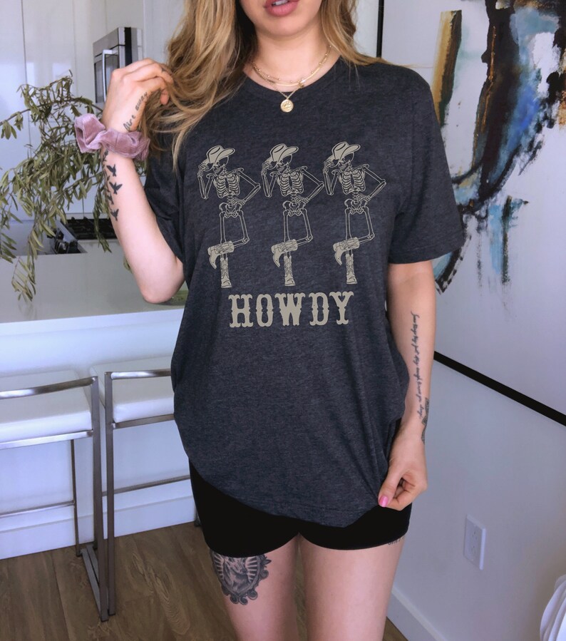 K&ouml;nnte beinhalten: Ein dunkelgraues T-Shirt mit einem Aufdruck von drei Skeletten, die Cowboyh&uuml;te und Stiefel tragen. Die Skelette stehen in einer Reihe und der Text "HOWDY" ist darunter gedruckt.