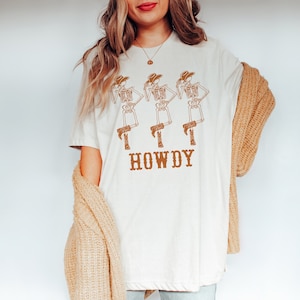 K&ouml;nnte beinhalten: Wei&szlig;es T-Shirt mit einem Aufdruck von drei Skeletten, die Cowboyh&uuml;te und Stiefel tragen. Der Text "HOWDY" ist unter den Skeletten gedruckt.