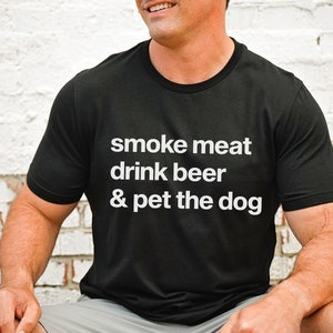 K&ouml;nnte beinhalten: Ein schwarzes T-Shirt mit wei&szlig;em Text, der "smoke meat drink beer & pet the dog" lautet.