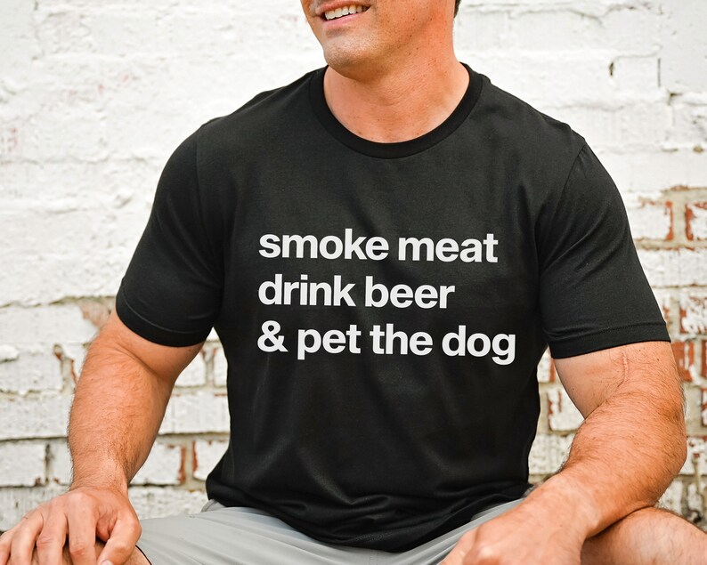 K&ouml;nnte beinhalten: Ein schwarzes T-Shirt mit wei&szlig;em Text, der "smoke meat drink beer & pet the dog" lautet.