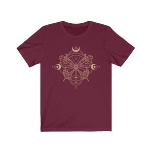 May include: A maroon t-shirt with a gold geometric design featuring a butterfly and crescent moons.
