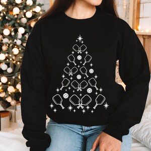 K&ouml;nnte beinhalten: Ein schwarzes Sweatshirt mit einem wei&szlig;en Design eines Weihnachtsbaums aus Pickleball-Schl&auml;gern und B&auml;llen.
