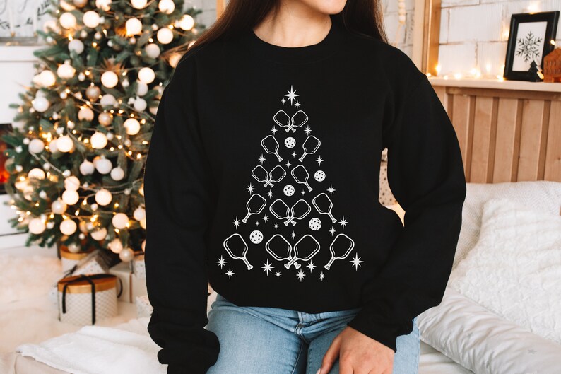 K&ouml;nnte beinhalten: Ein schwarzes Sweatshirt mit einem wei&szlig;en Design eines Weihnachtsbaums aus Pickleball-Schl&auml;gern und B&auml;llen.