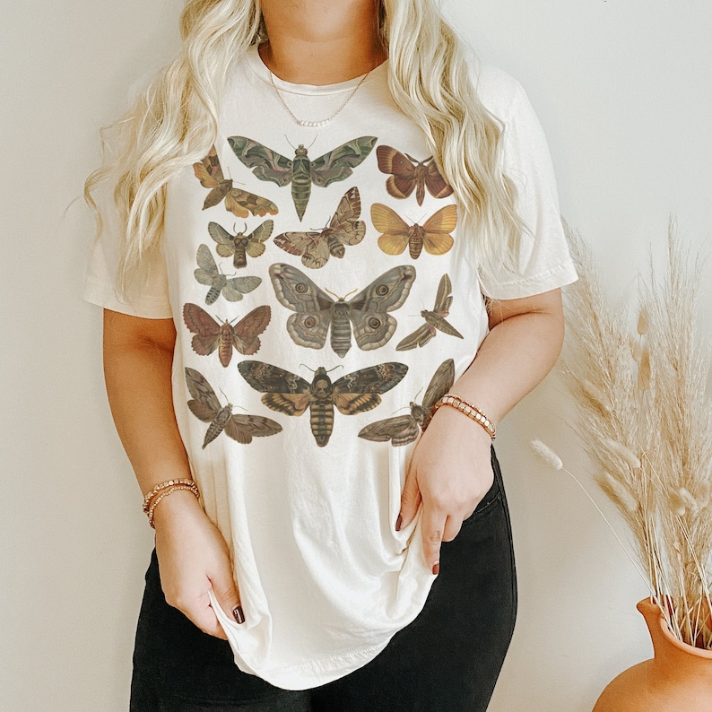May include: A white t-shirt with a graphic print of various butterflies and moths in shades of brown, green, and black. The shirt has a relaxed fit and a crew neckline.