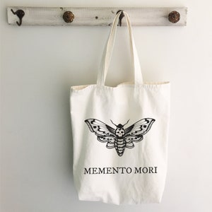 May include: A white canvas tote bag with a black and white illustration of a moth with a skull on its body. The text "MEMENTO MORI" is printed below the moth.