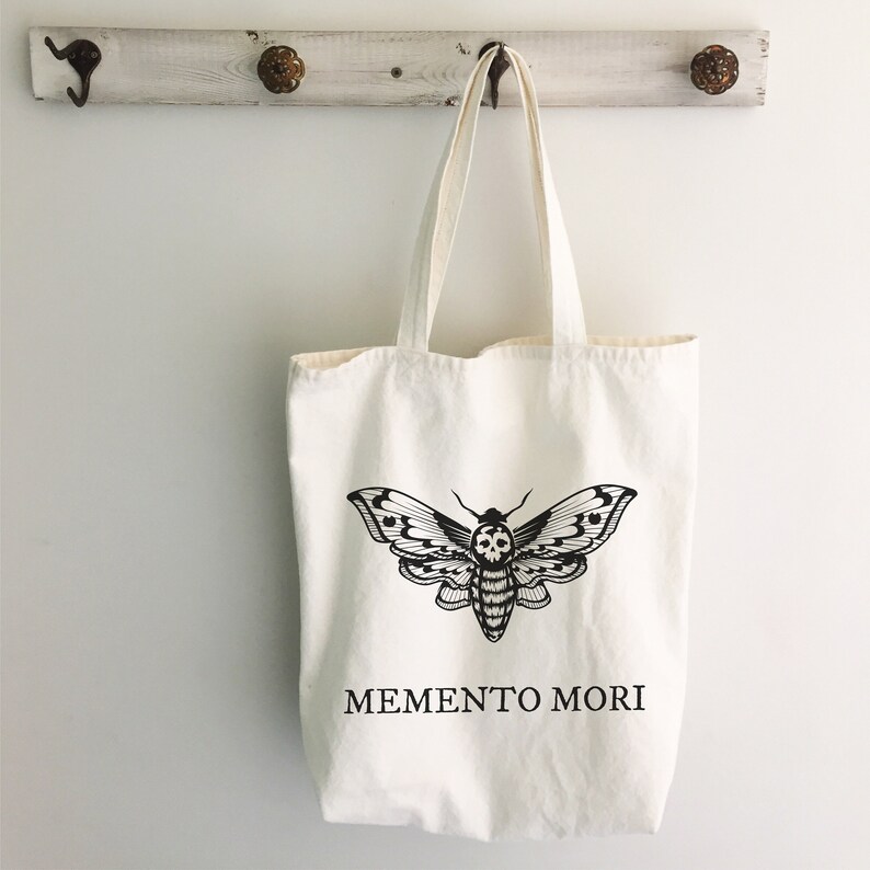 May include: A white canvas tote bag with a black and white illustration of a moth with a skull on its body. The text "MEMENTO MORI" is printed below the moth.