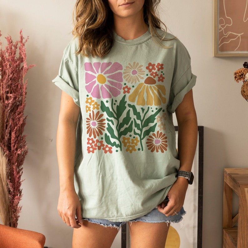 Puede incluir: Una camiseta verde claro con un dise&ntilde;o floral colorido. El gr&aacute;fico incluye flores rosas, amarillas y naranjas con hojas verdes. La camiseta tiene mangas enrolladas y un corte holgado.