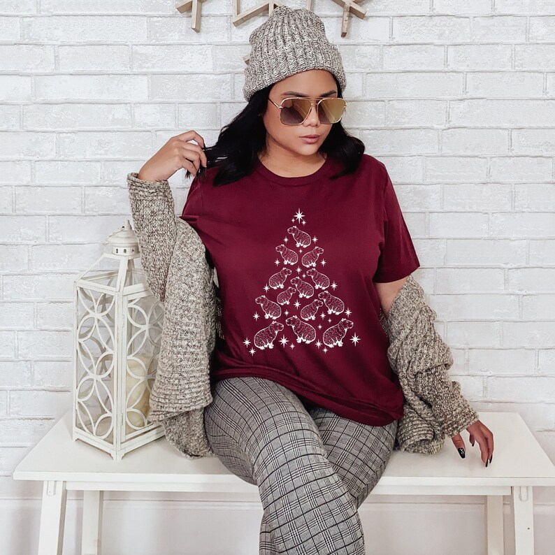 May include: A maroon t-shirt featuring a white outline of a Christmas tree made of sheep. The tree is decorated with a star at the top and has a white outline.