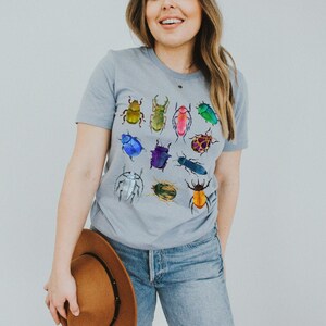 May include: A light gray t-shirt with a colorful beetle design. The beetles are in various shades of green, blue, red, yellow, and purple.