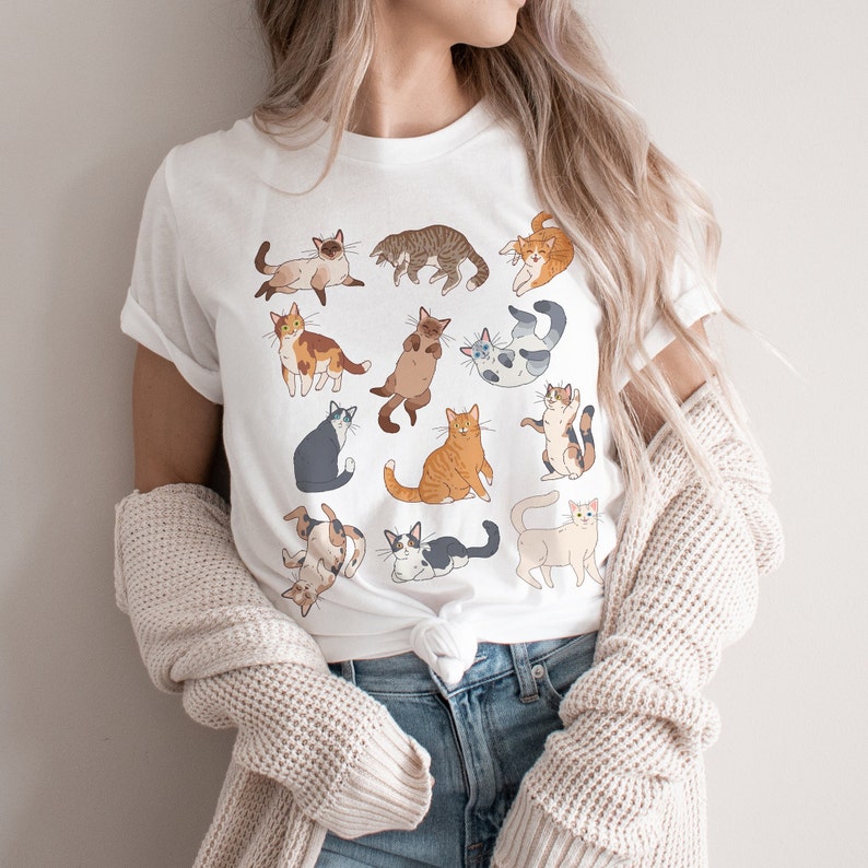 May include: White t-shirt featuring a collection of illustrated cats in various poses and colors. The cats are depicted in a playful and whimsical style, with a variety of patterns and expressions. The shirt is made of a soft, comfortable material.