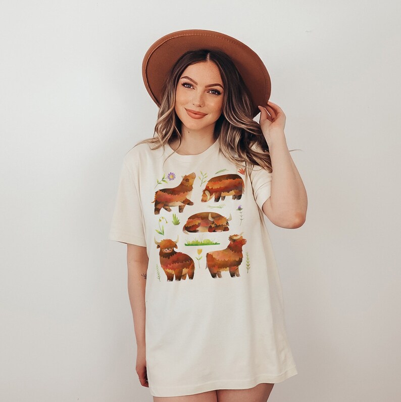 Puede incluir: Una mujer con una camiseta blanca con un dise&ntilde;o de vaca de las tierras altas de color marr&oacute;n, naranja y amarillo. La camiseta tiene cuello redondo y mangas cortas. La mujer lleva un sombrero marr&oacute;n y tiene el pelo largo y casta&ntilde;o.