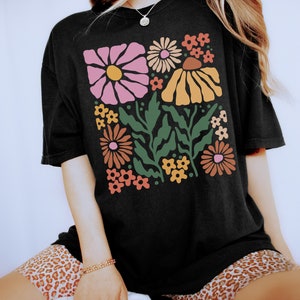Puede incluir: Una camiseta negra con un estampado floral retro con flores rosas, naranjas, amarillas y verdes. Las flores son estilizadas y tienen un toque caprichoso.