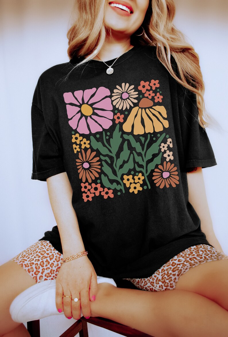 Puede incluir: Una camiseta negra con un estampado floral retro con flores rosas, naranjas, amarillas y verdes. Las flores son estilizadas y tienen un toque caprichoso.
