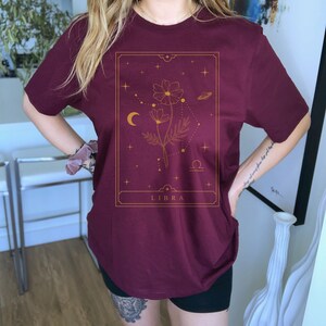 May include: A maroon t-shirt with a gold graphic of a flower, stars, a crescent moon, and the word "LIBRA" in a rectangle.