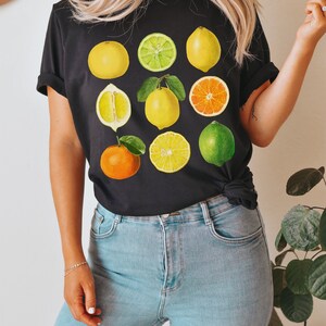 Pode incluir: Uma camiseta preta com uma estampa gr&aacute;fica de v&aacute;rias frutas c&iacute;tricas, incluindo lim&otilde;es, limas, laranjas e toranjas. As frutas est&atilde;o dispostas em uma grade na camiseta.