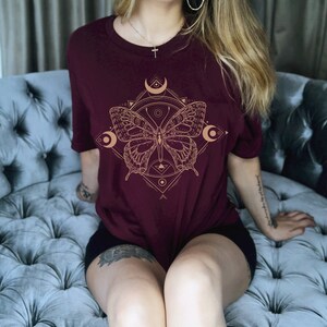 May include: A maroon t-shirt with a gold geometric design featuring a butterfly and crescent moons.