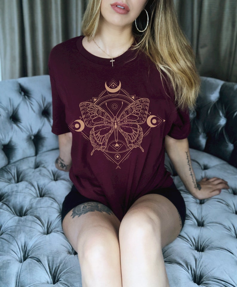 May include: A maroon t-shirt with a gold geometric design featuring a butterfly and crescent moons.