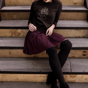 May include: A woman wearing a dark brown sweatshirt with a white line drawing of a face with leaves on top and the text "grow where you are planted". She is wearing a burgundy pleated skirt and black tights with black ankle boots. She is sitting on wooden stairs.