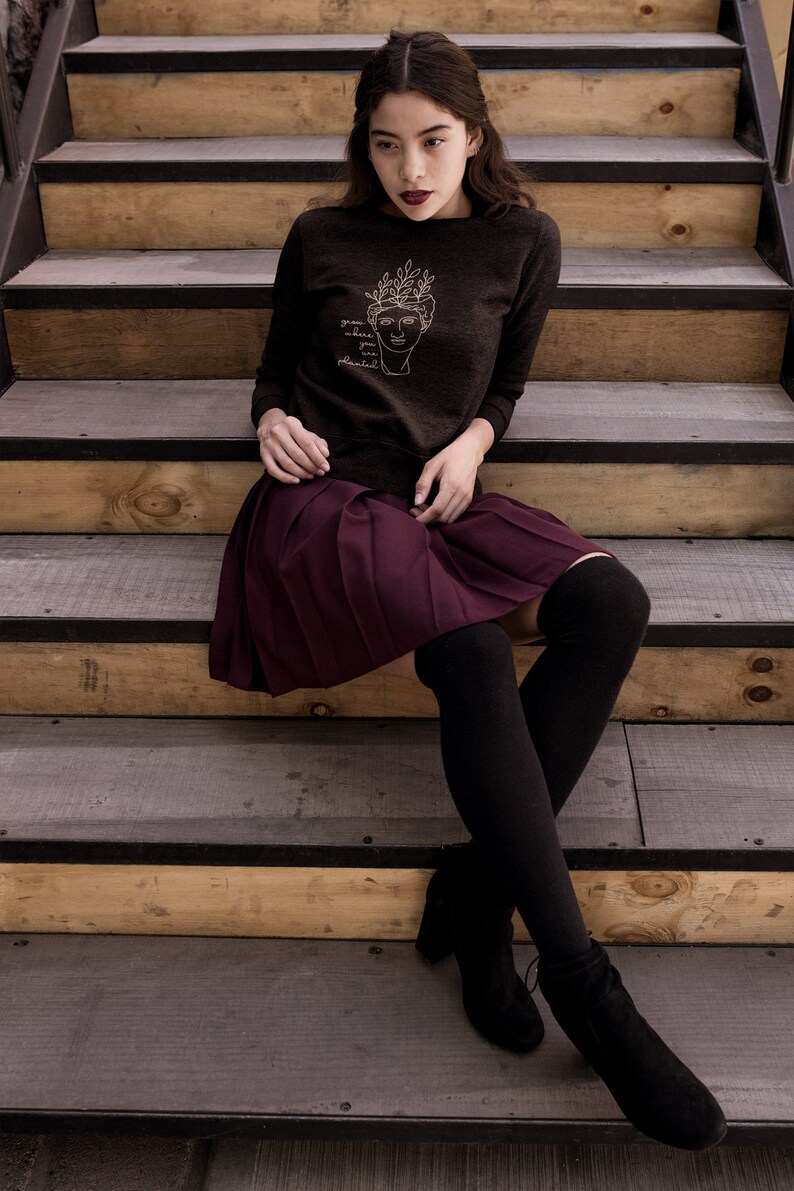 May include: A woman wearing a dark brown sweatshirt with a white line drawing of a face with leaves on top and the text "grow where you are planted". She is wearing a burgundy pleated skirt and black tights with black ankle boots. She is sitting on wooden stairs.