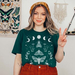 Puede incluir: Una mujer con una camiseta verde con un dise&ntilde;o gr&aacute;fico blanco de luna y polilla. La camiseta tiene un fondo verde oscuro y presenta una luna blanca, estrellas y polillas. La mujer sonr&iacute;e y lleva el pelo recogido en una diadema.