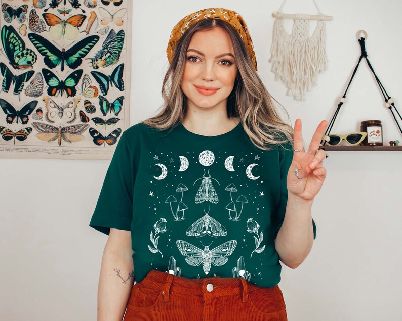 Puede incluir: Una mujer con una camiseta verde con un dise&ntilde;o gr&aacute;fico blanco de luna y polilla. La camiseta tiene un fondo verde oscuro y presenta una luna blanca, estrellas y polillas. La mujer sonr&iacute;e y lleva el pelo recogido en una diadema.