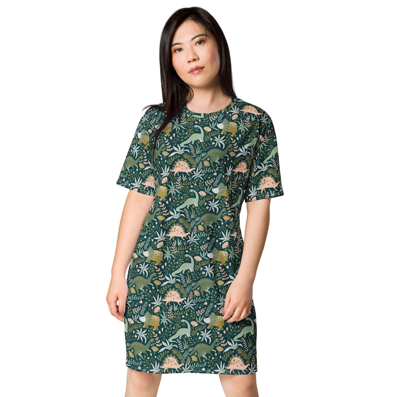 Op de afbeelding: Een groene t-shirtjurk met een dinosaurus- en bladmotief. De jurk heeft korte mouwen en een ronde hals.