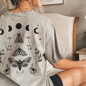 Op de afbeelding: Een grijs T-shirt met een grafische print van motten, paddenstoelen en manen. De grafiek is in zwart en wit.