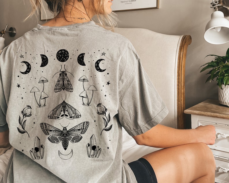 Op de afbeelding: Een grijs T-shirt met een grafische print van motten, paddenstoelen en manen. De grafiek is in zwart en wit.