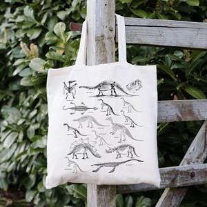 May include: A white canvas tote bag with a black and white illustration of dinosaur skeletons. The bag features a variety of dinosaur skeletons, including a Tyrannosaurus Rex, a Stegosaurus, and a Triceratops.