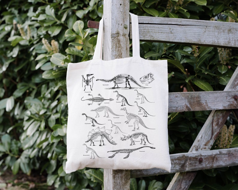 May include: A white canvas tote bag with a black and white illustration of dinosaur skeletons. The bag features a variety of dinosaur skeletons, including a Tyrannosaurus Rex, a Stegosaurus, and a Triceratops.